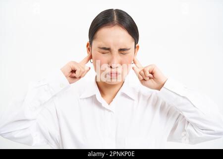 Infastidita giovane operatrice asiatica d'impresa, signora urla e chiude le orecchie con le dita, si trova frustrato contro sfondo bianco studio Foto Stock