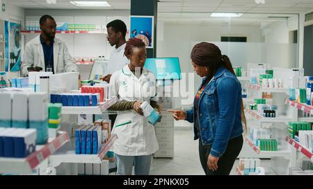 Farmacista femminile aiutare il cliente con la medicina, mostrando prodotti farmaceutici da scaffali farmacia. Giovane adulto che chiede specialista di salute su scatole di pillole e integratori. Foto Stock