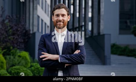 Uomo d'affari sicuro riuscito in vestito elegante che si erge sull'edificio dell'ufficio del fondo sorridente guardando la macchina fotografica il leader maschio caucasico bello Foto Stock