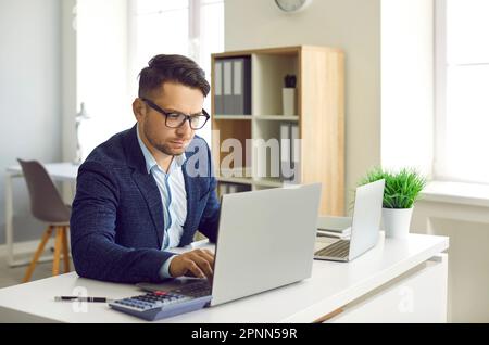 Uomo d'affari serio e occupato che lavora sul computer portatile e utilizza la calcolatrice per i calcoli. Foto Stock