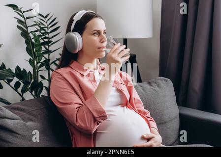 Ragazza incinta simpatica che ascolta la musica con le cuffie wireless, goditi l'acqua minerale fresca e pura al mattino. Futura madre tenere in vetro swallow Foto Stock