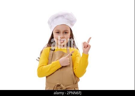 Sorridente ragazza felice del bambino che indossa il cappello del cuoco ed il grembiule beige della cucina, punta le barrette allo spazio dell'annuncio della copia sullo sfondo bianco Foto Stock