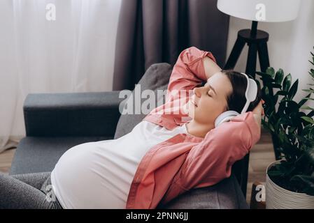 Felice giovane donna incinta indossando cuffie wireless ascoltare musica mentre sdraiato sul comodo divano nel soggiorno di casa, godere hea Foto Stock