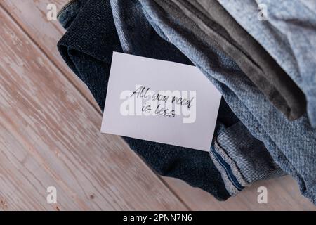 TUTTO CIÒ DI CUI HAI BISOGNO È MENO testo su carta nota su Jeans assortimento abbigliamento shopping sostenibile di seconda mano. Armadio a capsula minimal. Consumo eccessivo di moda sostenibile, consumo di acquisto cosciente, concetto di idea di modo lento. Stile di vita a basso contenuto di rifiuti Foto Stock