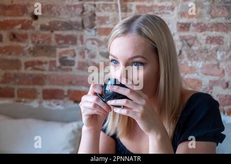 Ritratto di giovane donna gioiosa che si gode una tazza di caffè a casa. Bella ragazza sorridente che beve il tè caldo in inverno. Giovane donna bionda che indossa una blusa nera il giorno della primavera. Foto di alta qualità Foto Stock