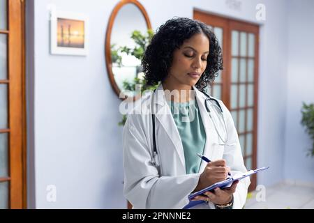 Medico di sesso femminile biraciale che indossa stetoscopio con la clipboard e prende appunti presso l'ufficio del medico Foto Stock