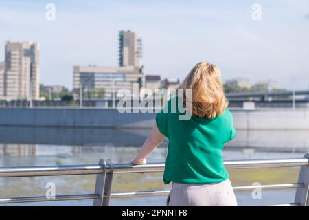 Una vista dal retro di una ragazza in una blusa verde in piedi sulla riva del fiume sullo sfondo di una città sfocata. Una ragazza cammina per le strade Foto Stock