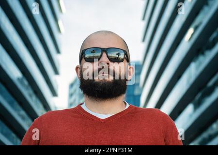 Ritratto di uomo barbuto freelancer o specialista IT in occhiali da sole con riflessi di palma e spiaggia dopo il lavoro al di fuori con moderni edifici di uffici su b Foto Stock