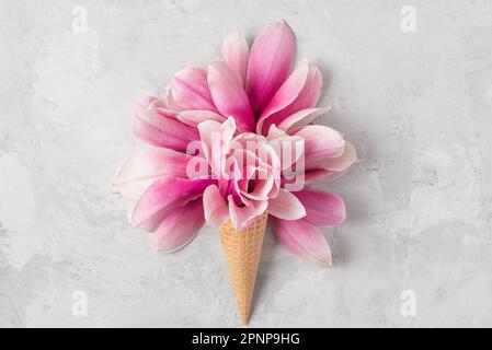 Cono gelato con fiori di magnolia rosa su fondo in cemento. Concetto di vacanza primaverile. Giacitura piatta. Vista dall'alto. Layout creativo Foto Stock