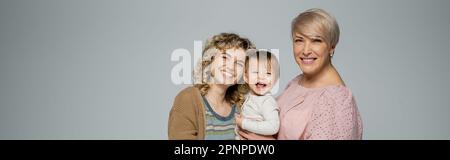 ragazza bambino eccitato ridendo vicino a madre felice e nonna isolato su grigio, banner, immagine stock Foto Stock