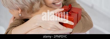 vista parziale di donna sorridente con scatola regalo rossa che abbraccia mamma di mezza età a casa, banner, immagine stock Foto Stock