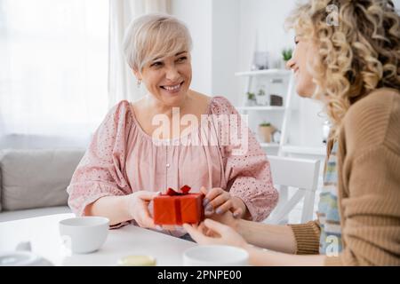 felice donna di mezza età che tiene presente e guardando la figlia adulta mentre si siede vicino a tazze da tè in cucina, immagine stock Foto Stock