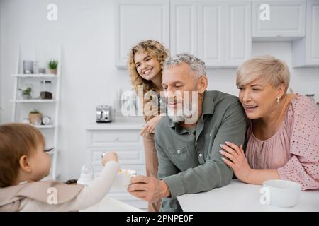 uomo sorridente che tiene la ciotola mentre mangina il bambino con la colazione vicino alla famiglia in cucina, immagine di scorta Foto Stock