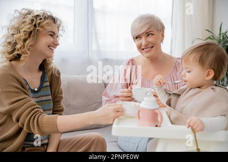 felice donna matura con tè tazza guardando figlia adulto che alimenta bambina in cucina, immagine stock Foto Stock