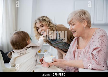 bambina che alimenta mamma vicino a nonna sorridente con ciotola in cucina, immagine stock Foto Stock