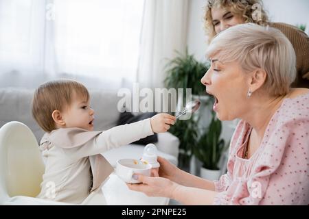 vista laterale del bambino piccolo con cucchiaio che alimenta la nonna con il recipiente in mani, immagine di scorta Foto Stock