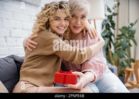 felice donna matura con figlia adulto abbracciando e guardando la macchina fotografica vicino al contenitore del regalo in soggiorno, immagine di scorta Foto Stock