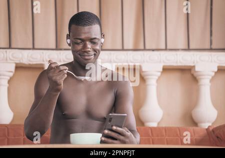 Content l'uomo afroamericano mangia porridge e legge le notizie sul cellulare durante la colazione a casa. L'immagine cattura le sue capacità di multitasking e re Foto Stock