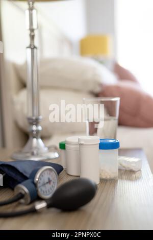 Primo piano dei medicinali, monitor della pressione sanguigna e bicchiere d'acqua sul comodino in camera da letto Foto Stock