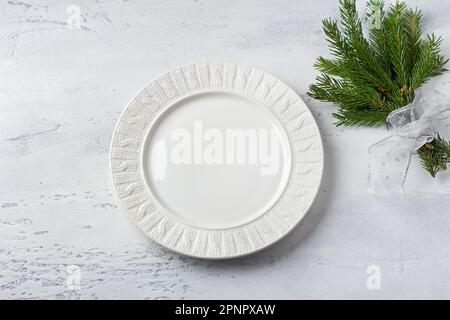 Piatto di ceramica bianca con rami di albero di natale decorato con nastro d'argento su sfondo grigio chiaro testurizzato, vista dall'alto Foto Stock