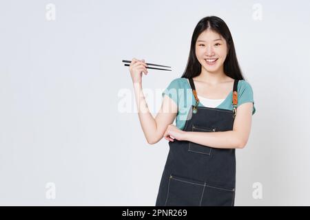 Giovane donna asiatica che indossa grembiule da cucina con bacchette isolate su sfondo bianco. Foto Stock