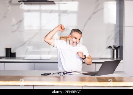 Uomo eccitato in bicchieri sedersi a tavola in cucina sentire euforico vincere lotteria online sul computer portatile, overjoyed giovane maschio in occhiali guardare lo schermo del computer Foto Stock