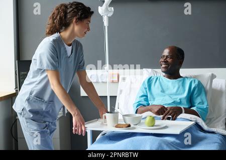 Giovane infermiera in uniforme che porta il pranzo per il paziente anziano mentre si siede a letto nel reparto di ospedale Foto Stock