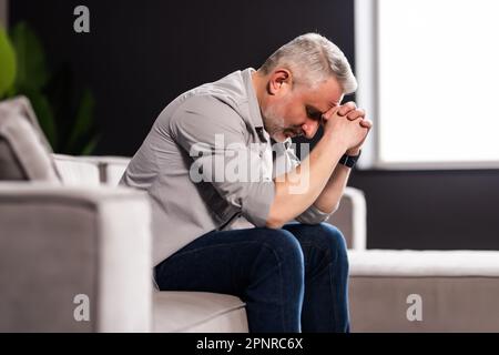 Frustrato uomo di mezza età seduto sul divano a casa. Primo piano faccia di un uomo d'affari stressato che indossa gli occhiali con gli occhi chiusi. Iperlavorato medio-est Foto Stock