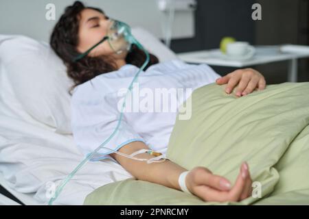 Paziente giovane con catetere nel braccio e maschera per ossigeno sdraiata sul letto nel reparto ospedaliero Foto Stock