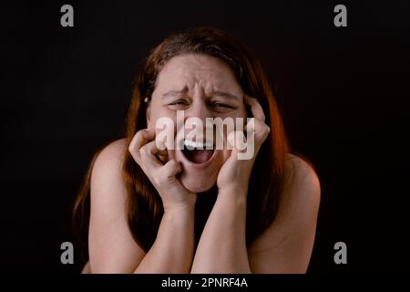 Giovane donna piangente che graffia il viso. Grida e grida forte a bocca aperta. Sentimenti, dolore, sofferenza. Emozioni umane. Foto Stock