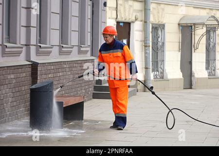 Donna in uniforme operaio innaffiare la panca con un tubo flessibile. Pulizia della strada e disinfezione nella città di primavera Foto Stock