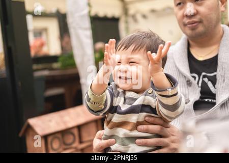 Bambino maschio sorridente alzando le mani nell'eccitamento seduto sulle gambe del suo padre, il papà sta tenendo il bambino con le sue mani. All'aperto in una birreria all'aperto in PU Foto Stock