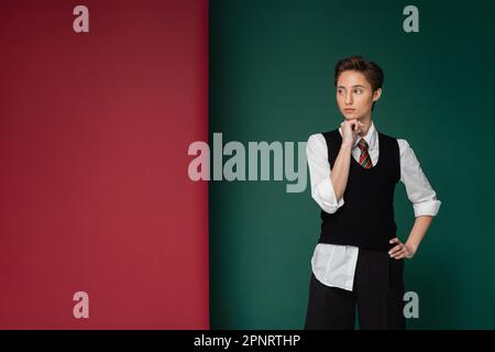 elegante giovane studente con capelli corti in posa con mano sull'anca su sfondo verde e marrone, immagine stock Foto Stock