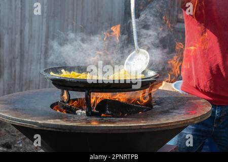 Le verdure sono tostate su un focolare rotondo. Griglia rotonda a forma di ciotola con un fuoco all'interno. L'uomo prepara il focolare per il barbecue. La legna da ardere sta bruciando Foto Stock