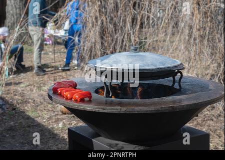 Le verdure sono tostate su un focolare rotondo. Griglia rotonda a forma di ciotola con un fuoco all'interno. L'uomo prepara il focolare per il barbecue. La legna da ardere sta bruciando Foto Stock