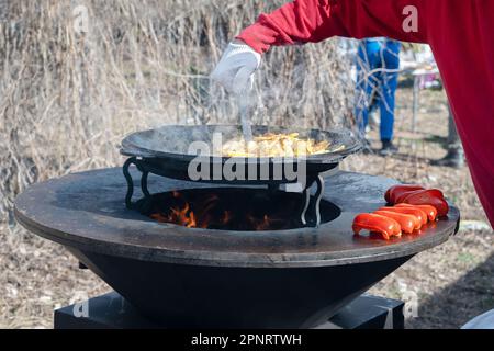 Le verdure sono tostate su un focolare rotondo. Griglia rotonda a forma di ciotola con un fuoco all'interno. L'uomo prepara il focolare per il barbecue. La legna da ardere sta bruciando Foto Stock