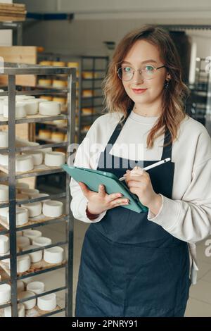 Young Farm Female Owner controlla il processo di maturazione delle teste di formaggio di capra poste su scaffali in magazzino a Cheese Factory. Scrittura di note con Digital T. Foto Stock