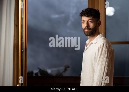 Un giovane uomo dai capelli neri ricci-capelli si alza di sera ad una porta aperta contro un bellissimo sfondo serale Foto Stock