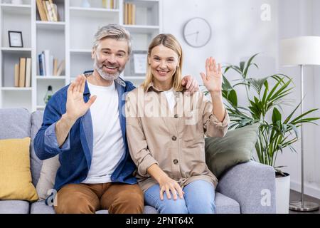 Felice coppia di famiglia uomo e donna maturi seduti insieme sul divano in soggiorno e di scambio guardando la fotocamera sorridente e la mano agitando saluto gesto, videochiamata webcam vista. Foto Stock