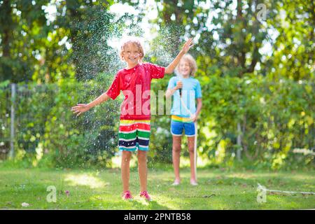 I bambini giocano con il tubo di spruzzatura dell'acqua. Estate giardino all'aperto divertimento per i bambini. Il ragazzo spruzzi d'acqua nelle calde giornate di sole. Piante di annaffiamento di capretto in cortile. Foto Stock