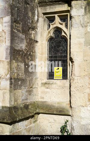 Avviso CCTV giallo visualizzato nella finestra con piombo di una vecchia chiesa in pietra protetta da una griglia metallica Foto Stock