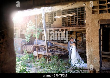 Sposa e sposo tenendo mani e abiti da sposa baciare in vecchia fabbrica di mattoni abbandonati con raggio di sole in un angolo che illumina la scena Foto Stock