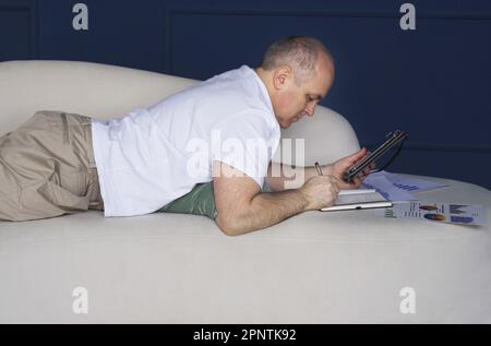 Un uomo d'affari si trova su un divano e guarda le tendenze dei dati su grafici e grafici in un tablet, prende appunti in un notebook. Lavoro a distanza Foto Stock