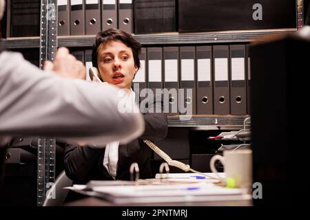 Donna poliziotto che parla con l'ispettore remoto al telefono fisso, discutendo il rapporto delle indagini penali. I detective privati lavorano a tarda notte in una stanza arhive, analizzando i file delle prove Foto Stock