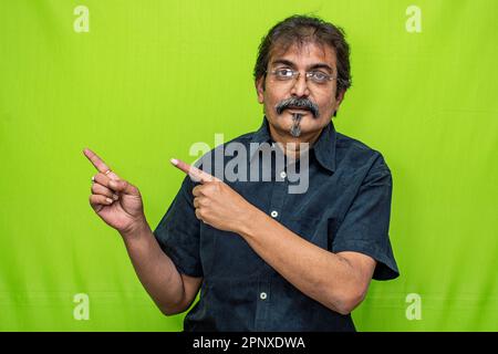 L'astuto uomo aziendale in camicia nera e spettacolo indica la giusta direzione con entrambe le dita indice, in piedi contro uno sfondo verde schermo Foto Stock