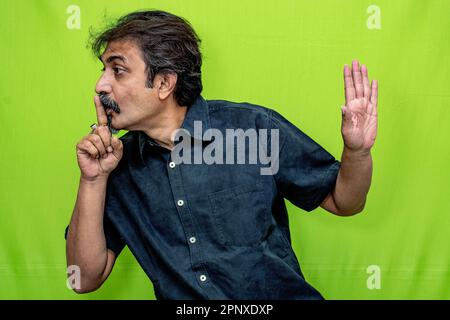 Un uomo corporativo in camicia nera, in piedi contro uno schermo verde, segnala silenzio con il dito indice destro sulle labbra e gesti con la mano sinistra a sinistra Foto Stock