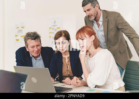 Un gruppo diversificato di quattro colleghi senior e junior si riunisce intorno a un computer portatile, studiando un progetto insieme a espressioni focalizzate, vestito in smart ca Foto Stock