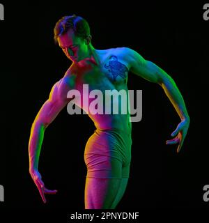 Ritratto di ballerina maschile muscolosa e aggraziata che balla sullo sfondo scuro dello studio con luce al neon. Vista posteriore. Attore flessibile e senza peso Foto Stock