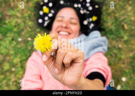 Donna sorridente sdraiata sull'erba mostrando un dente di leone nelle sue mani, rilassata e divertente. Concetto di primavera relax e connessione con la natura. Vista dall'alto. Foto Stock