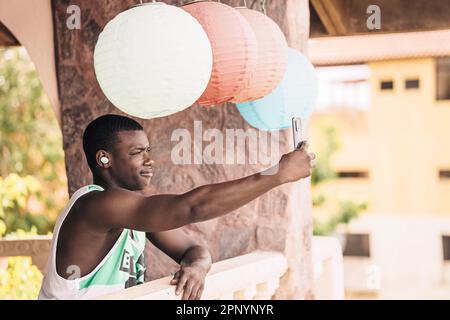 Vista laterale del bel giovane uomo nero che indossa un canotta e sorride mentre prende un selfie con grandi ornamenti di carta sulla sua testa su una terrazza dell'hotel. Foto Stock